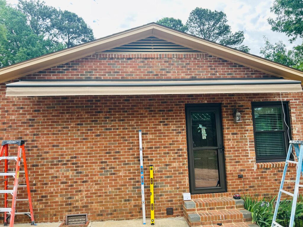 A brick house with a tan awning—installed by a trusted SunPro dealer—shades a black door and window. Two ladders and two measuring tools lean against the wall, ready for further work. Green trees are visible above the roofline.