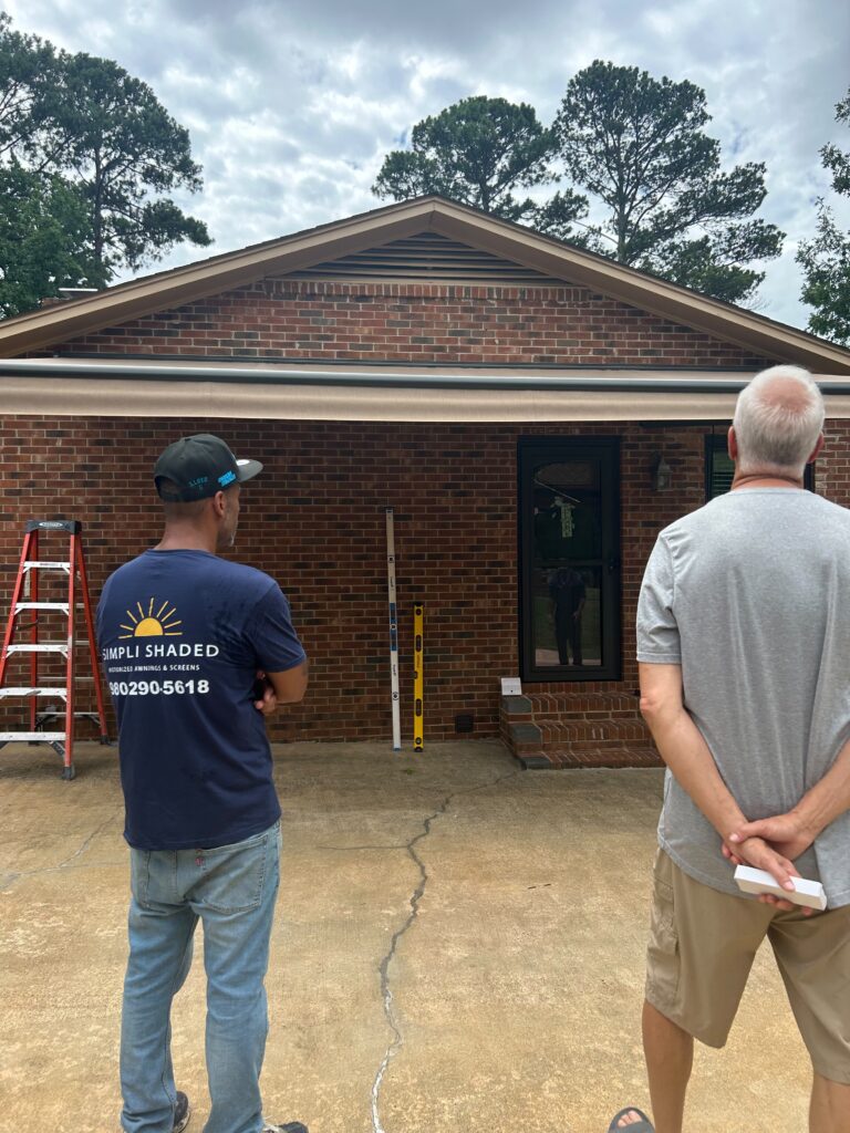 Two men stand in front of a brick house with a garage—one wears a blue shirt and cap, the other a gray shirt. A red ladder and measuring tools, essential for any Sunpro dealer, are nearby. Trees and a cloudy sky form the backdrop.