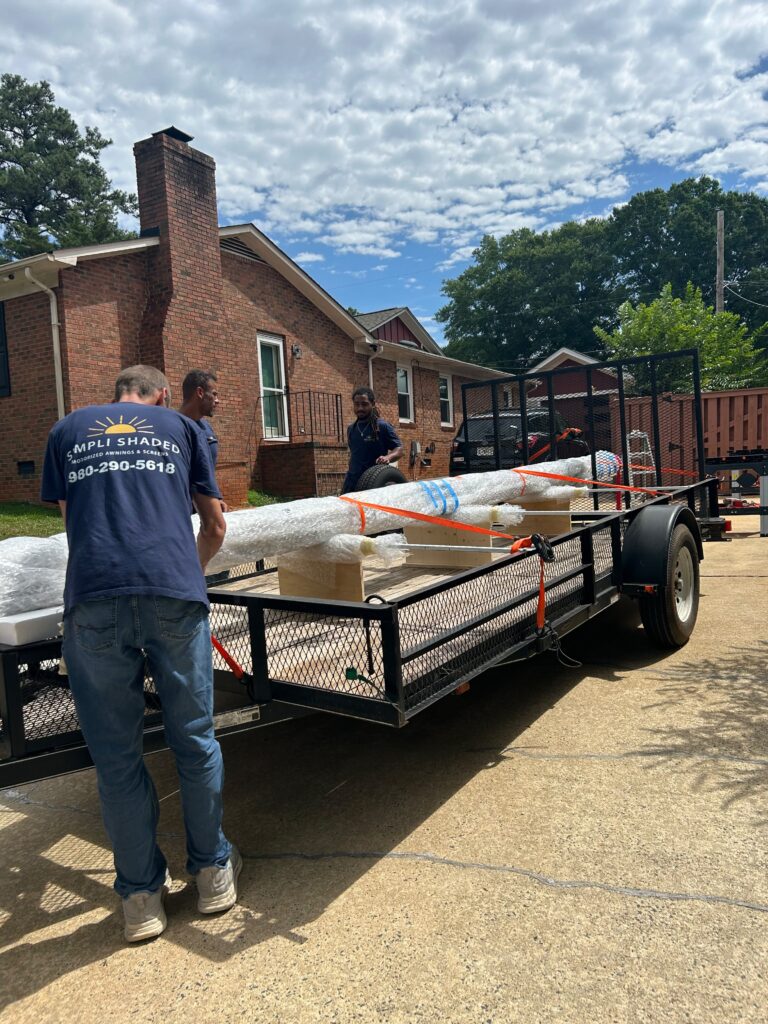 Three workers, possibly from a SunPro dealer, load or unload wrapped items from a flatbed trailer parked in a residential driveway. A brick house and trees are in the background under a partly cloudy sky.